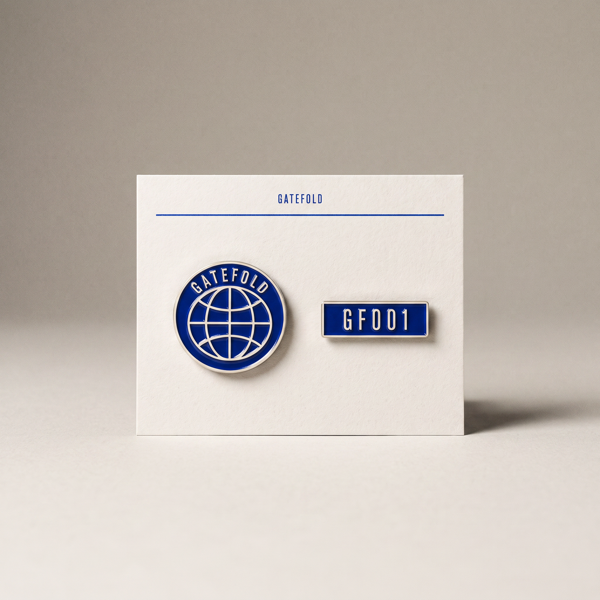 Two cobalt enamel pins on an off-white card: the GATEFOLD globe roundel and a GF001 bar